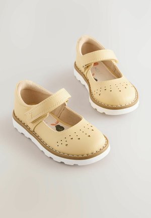 Chaussures Mary Jane en cuir jaune avec une bride velcro, des cœurs perforés sur la zone des orteils et une semelle en caoutchouc texturé blanche.