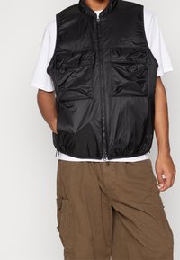 Svart lättviktig väst med framdragkedja, hög krage och två bröstfickor. Bärs över en vit T-shirt, tillsammans med bruna cargohorts.