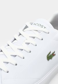 Adidași albi cu șireturi, cu un brad din piele netedă, limbă din material textil și un logo brodat în verde în formă de crocodil pe lateral.