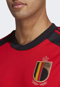 Camiseta deportiva roja con cuello negro, adornada con el escudo de la Real Asociación Belga de Fútbol en dorado y franjas de colores rojo, amarillo y negro.