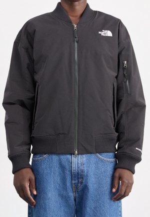 Personne portant une veste bomber noire North Face fermée par une fermeture éclair, associée à un jean bleu sur un fond uni.