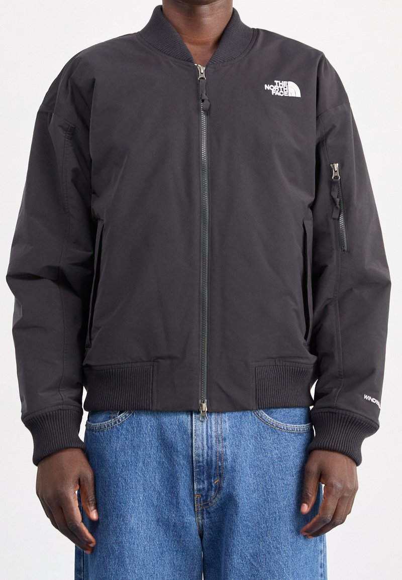 Personne portant une veste bomber noire North Face fermée par une fermeture éclair, associée à un jean bleu sur un fond uni.