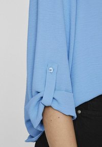 Blusa azzurra con maniche arrotolate, dotata di una chiusura a singolo bottone. Il tessuto è testurizzato e abbinato a pantaloni scuri.