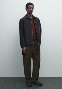 Black zip-up jacket over a brown sweater, paired with dark brown wide-leg trousers and dark shoes, standing on a textured gray surface.