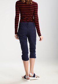 Námořnické capri kalhoty vyrobené z lehkého materiálu, spárované s dlouhým rukávem topem s černými a červenými pruhy. Bílé sandály se stříbrnými akcenty.