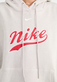 Světlě šedá mikina s velkým červeným logem "Nike" s puntíky. Obsahuje šňůrky na zavazování a žebrované manžety. Měkká texturovaná látka.