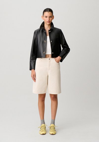 Mujer con chaqueta de cuero negra recortada, top corto blanco, pantalones cortos beige hasta la rodilla, calcetines grises y zapatillas amarillas, posando frente a un fondo liso.