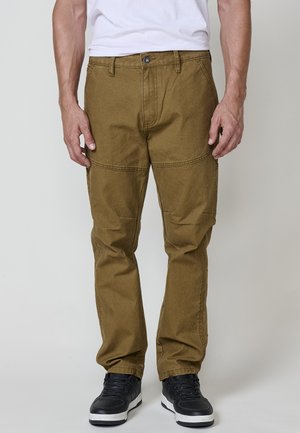 Pantalons cargo khaki avec une coupe droite, poches avant doubles et coutures renforcées. Fabriqués en tissu texturé, associés à des baskets noires.