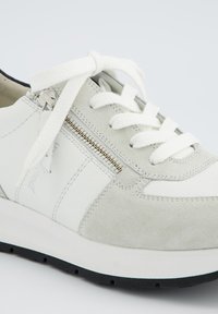 Sneaker blanche et grise avec un dessus en daim texturé et en cuir, dotée d'une fermeture éclair sur le côté, de lacets plats blancs et d'une semelle en caoutchouc noire.