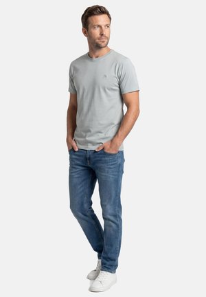 Man wearing light gray short-sleeve t-shirt, blue jeans, and white sneakers, standing with hands in pockets looking right.