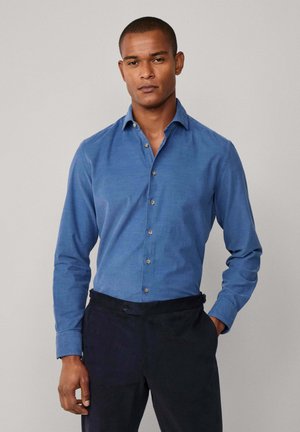Camisa de botones de color azul claro, hecha de una tela con una textura sutil. Presenta un collar clásico y mangas largas, combinada con pantalones oscuros.