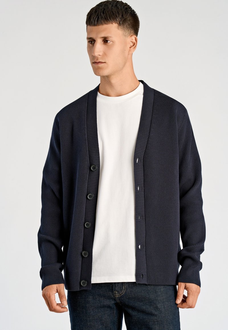Granatowy prążkowany kardigan z kołnierzem szalowym, zapinany z przodu na guziki, z długimi rękawami, noszony na białej koszuli, prezentujący luźny krój i teksturowaną tkaninę.