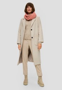 Long manteau beige avec des boutons noirs, superposé sur un pull léger beige et un pantalon beige. Présente une écharpe tricotée rose et des bottines beige.