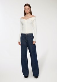 Biały sweter z dzianiny, z dekoltem off-the-shoulder, z zamkiem błyskawicznym z przodu, w połączeniu z luźnymi, ciemnoniebieskimi dżinsami. Gładka faktura tkaniny.