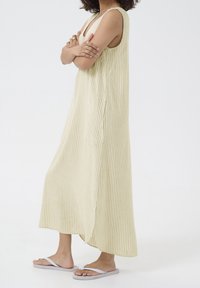 Robe maxi jaune clair à rayures blanches verticales, avec un décolleté en V profond et des fentes latérales, portée avec des tongs blanches.