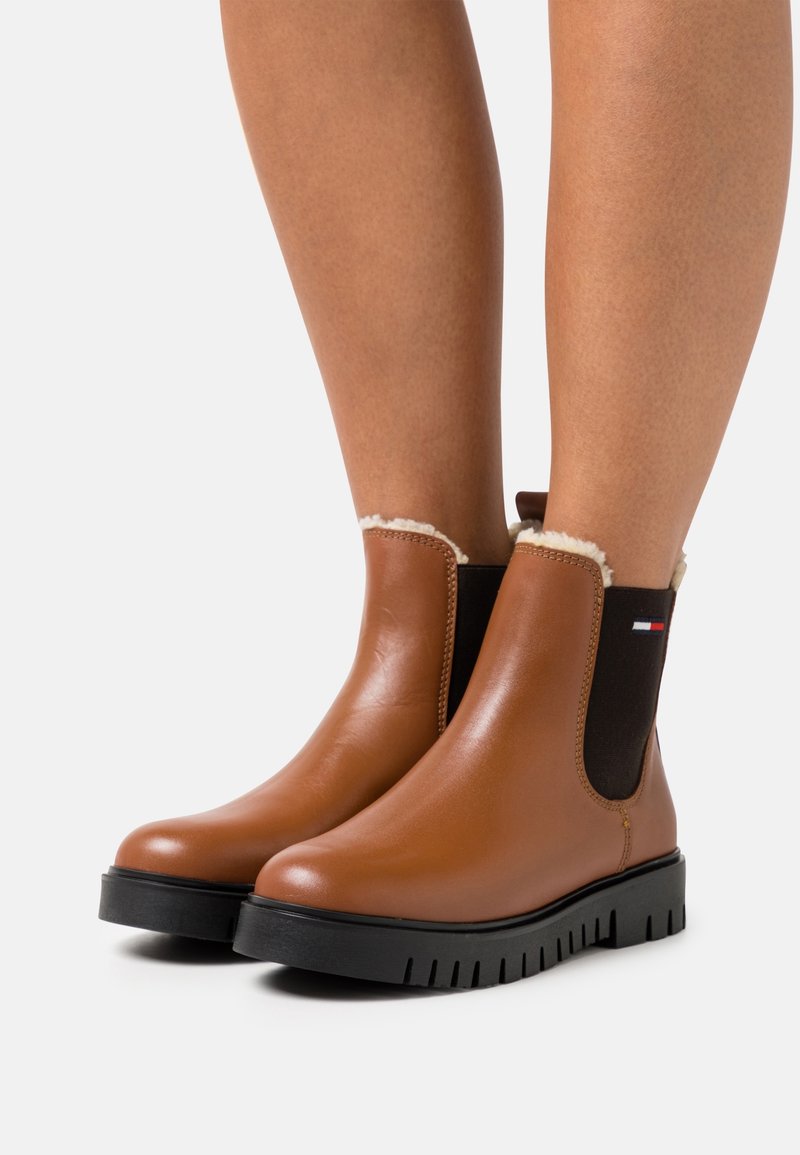 Bottes en cuir marron montant à la cheville avec panneaux latéraux élastiques noirs, doublure en polaire blanche, semelles épaisses à crampons noirs, portées sur des jambes nues.
