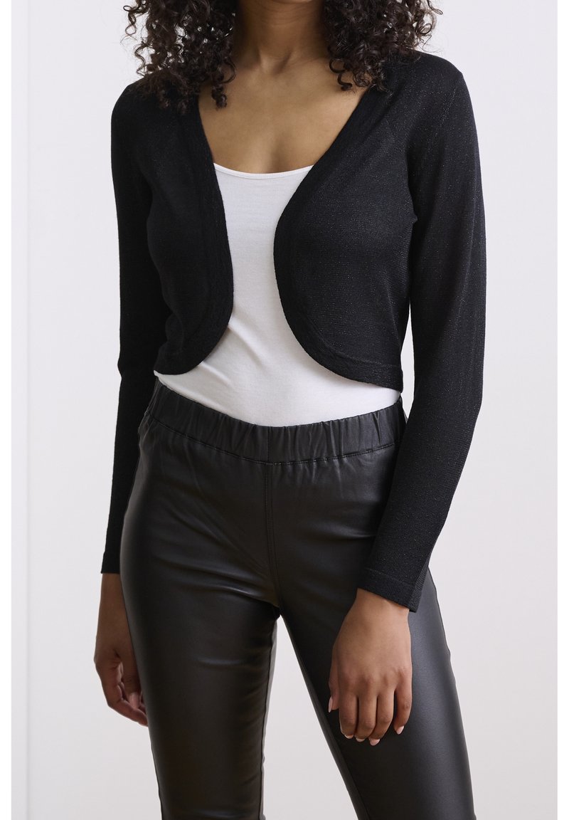 Schwarze, kurz geschnittene Strickjacke mit langen Ärmeln, aus einem leicht glänzenden Strickstoff, getragen über einem weißen Oberteil und kombiniert mit schwarzen, eng anliegenden Hosen.
