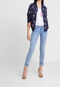 Kobieta ubrana w granatową bomberkę w kwiaty, białą koszulkę z grafiką, jasnoniebieskie obcisłe dżinsy z podwiniętymi nogawkami i cieliste sandały na wysokim obcasie.