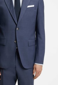 Homme portant un costume bleu foncé avec deux boutons fermés, une chemise blanche, un mouchoir blanc dans la poche et une cravate bleu marine à petits pois blancs.