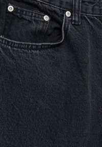Los jeans de mezclilla negros presentan un acabado texturizado, botones de metal y un bolsillo frontal. La tela tiene un ligero brillo y costuras visibles.
