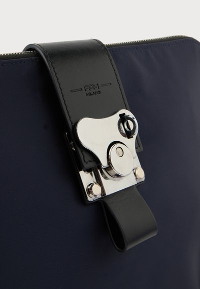 Gros plan d'un sac en tissu bleu marine avec une sangle en cuir noire et une boucle en métal argenté gravée avec "FPM Milano."
