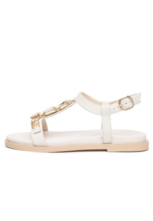 Weiße flache Sandale mit beiger Sohle, Knöchelriemen, goldener Schnalle und dekorativen goldenen sowie klaren Edelsteinverzierungen am Vorderriemen.