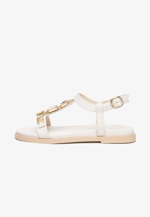 Sandale plate blanche avec semelle beige, bride de cheville, boucle dorée et ornements décoratifs en pierres précieuses dorées et transparentes sur la bride avant.