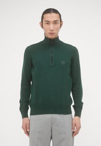 Groene zip-neksweater van fijne breimateriaal, met een geribbelde kraag, mouwen en zoom, en een klein logo op de borst.