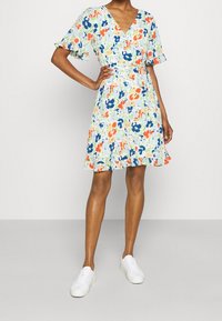 Květinové šaty s krátkým rukávem, které mají výstřih do V, stahovaný pas a sukni ke kolenům v kombinaci zelených, modrých, oranžových a bílých vzorů.