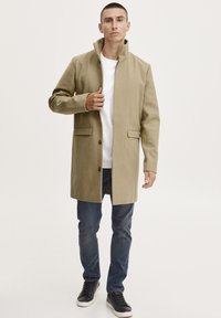 Cappotto beige con colletto alto, chiusura frontale con bottoni, due tasche laterali, indossato su una t-shirt bianca e jeans blu, abbinato a sneakers nere.