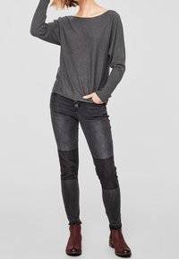 Grijze lange mouwen top met brede halslijn, gecombineerd met strakke zwarte jeans met paneeldetails. Draagt met donkerrode enkellaarzen.