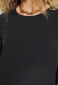 Musta paita, jossa on sileä pinta, pyöreä kaula-aukko ja lyhyet hihat, yhdistettynä kultaketjuun, jossa on geometrisiä linkkejä.