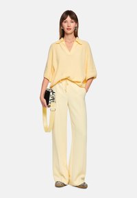 Light yellow knit sweater with a polo collar, paired with cream wide-leg trousers. Holds a patterned bag with a yellow strap.