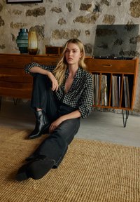 Melnas un baltas pārbaudītas krekls, melnas augstas vidukļa džinsus, norādītas melnas potītes zābaki; sēžot uz austā paklāja stilizētā interjerā.