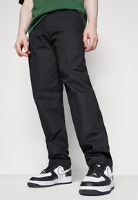 Svarta byxor med en slät yta och rak benmodell, kombinerat med svartvita sneakers med distinkta swoosh-detajler.