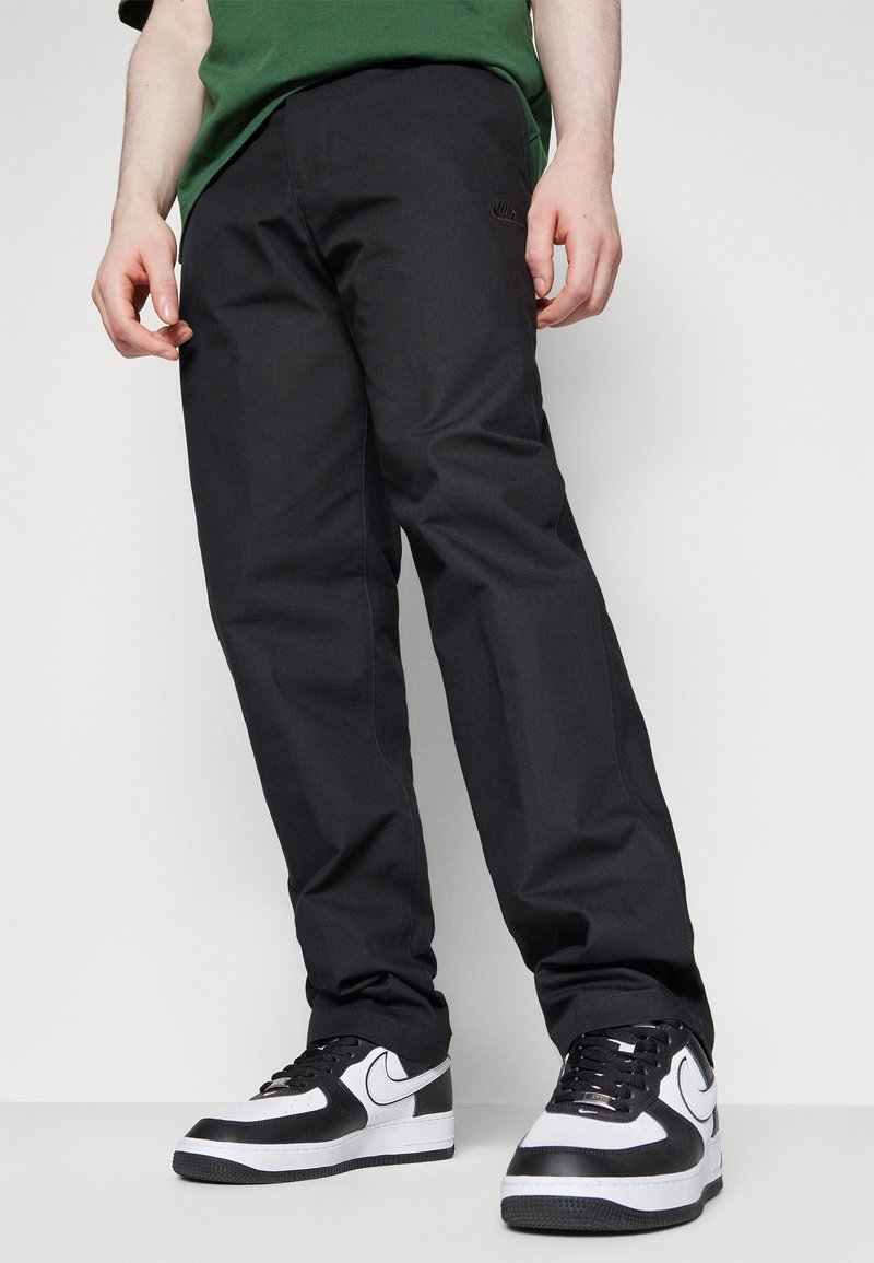Pantalons noirs avec une texture lisse et une coupe droite, associés à des baskets noir et blanc arborant des détails de swoosh distinctifs.