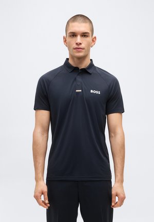 Νέος άνδρας με κοντά μαλλιά φοράει μαύρο πόλο με κοντά μανίκια και μικρό λογότυπο "BOSS" στο στήθος, στέκεται μπροστά σε απλό φωτεινό φόντο.