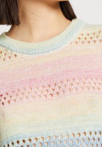 Sweter robiony na drutach z pastelowymi paskami w odcieniach różu, niebieskiego i zielonego, z ażurowymi detalami i ściągaczem przy dekolcie. Miękka, lekka tekstura.