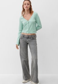 Muntgroene kort jasje met lange mouwen, V-hals en gekartelde zoom; gecombineerd met lichtgrijze flared jeans en crèmekleurige sneakers.