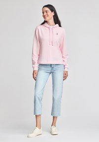 Rosa sweatshirt med huva, snörning och liten logotyp på bröstet, parat med ljusblåcroppade jeans och krämfärgade sneakers. Slät yta.