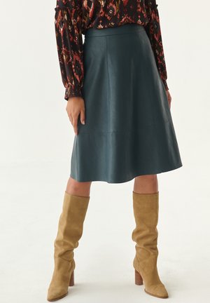 Jupe en cuir turquoise en forme de A avec une texture lisse, associée à des bottes en suède beige jusqu'aux genoux avec un talon carré. Motifs visibles sur le blouse.
