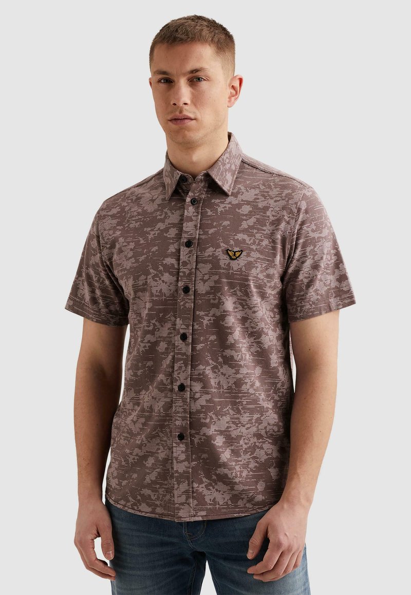 Jeune homme aux cheveux courts portant une chemise à manches courtes marron à motifs, boutonnée sur le devant, avec un petit emblème brodé sur la poitrine.