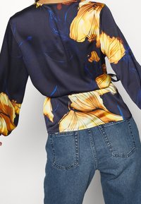 Bluse mit Blumenmuster und langen Ärmeln, navy Hintergrund, gelbe Blumen und einem gebundenen Taillengürtel, kombiniert mit blauen hochgeschnittenen Jeans.