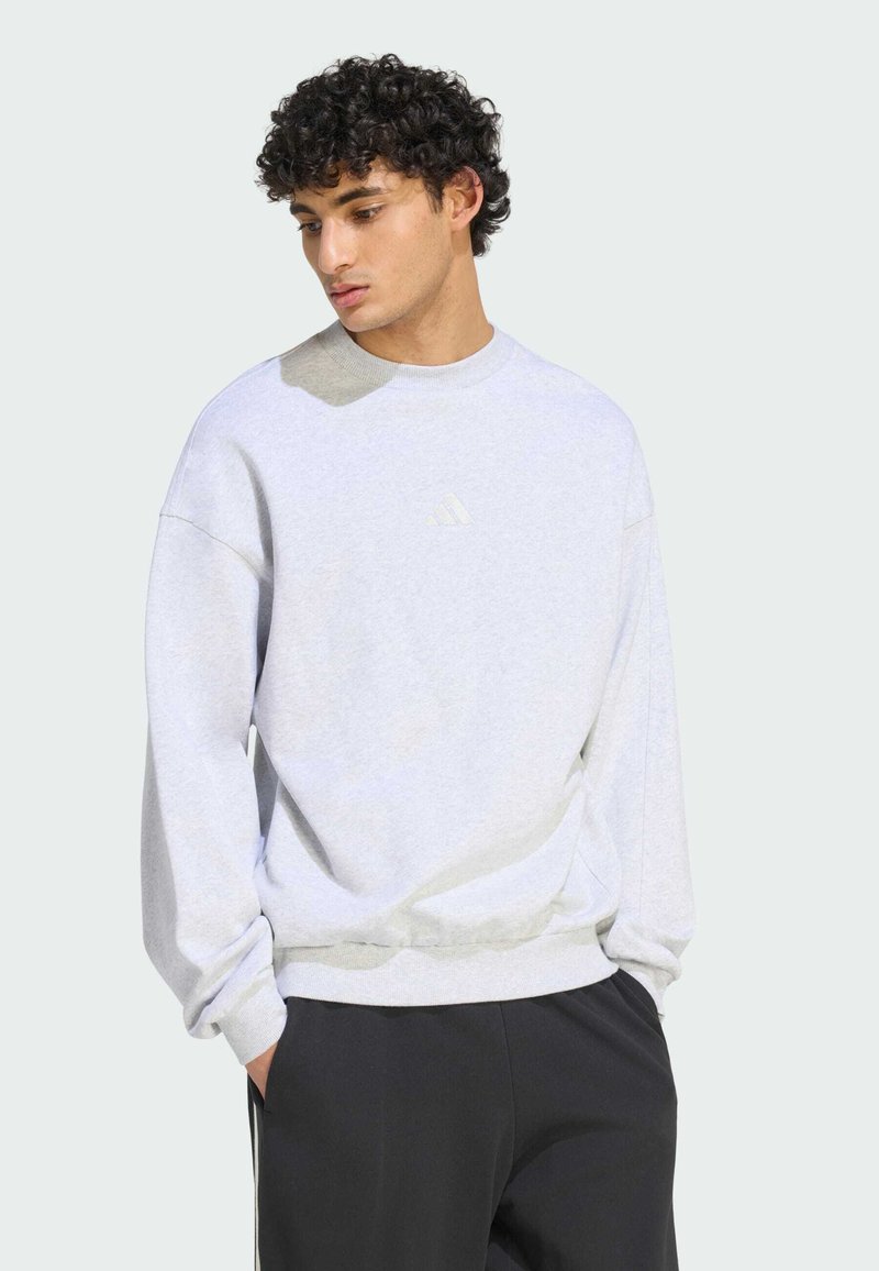 Jeune homme aux cheveux bouclés portant un sweat-shirt gris clair et un pantalon noir, debout les mains dans les poches, regardant vers le bas.