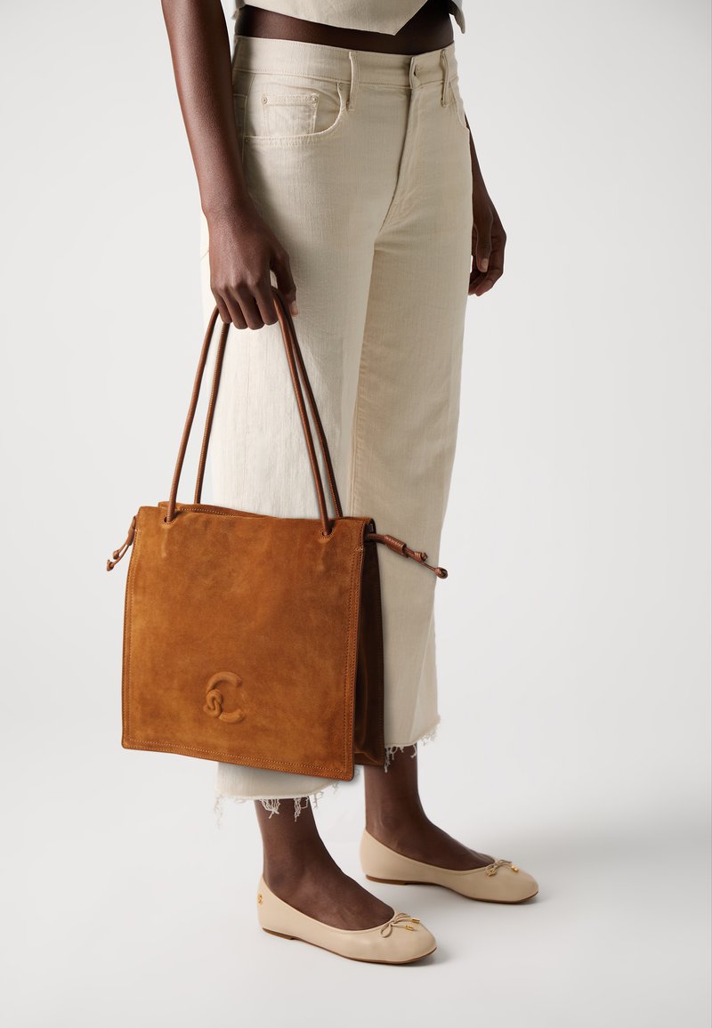 Person som holder en stor brun semsket lær tote bag med doble håndtak, iført kremfargede capribukser og beige ballerinasko mot en hvit bakgrunn.