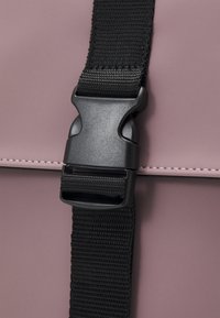 Detalle de bolso sintético rosa con una correa de nailon negra y hebilla de clip de plástico, con costuras en contraste a lo largo de los bordes.