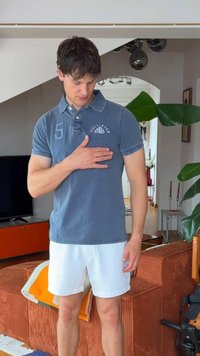Jeune homme en polo bleu et short blanc touche sa poitrine en se tenant dans un salon avec des plantes et des meubles.