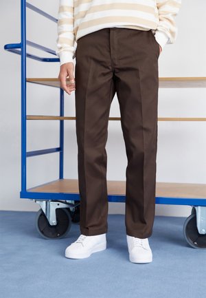 Braune Hose mit geradem Bein, die eine glatte Textur aufweist. Getragen mit einem gestreiften, cremefarbenen und beigen Oberteil sowie weißen Sneakern.