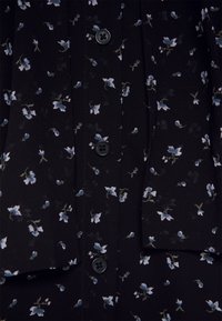 Close-up van zwarte stof met een klein wit en lichtblauw bloemmotief, met zwarte knopen voorzien van "Michael Kors" branding.