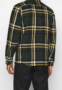 Chemise en flanelle à carreaux verts et noirs avec des accents jaunes et blancs, poignets boutonnés et col, vue de dos.